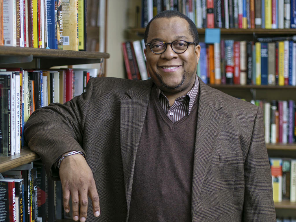 an african american male professor leaning on bookshelves with circular glasses