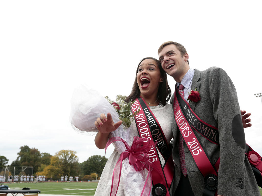 A male and female smiling proudly at a different camera, holding sashes and flowers to celebrate their victory