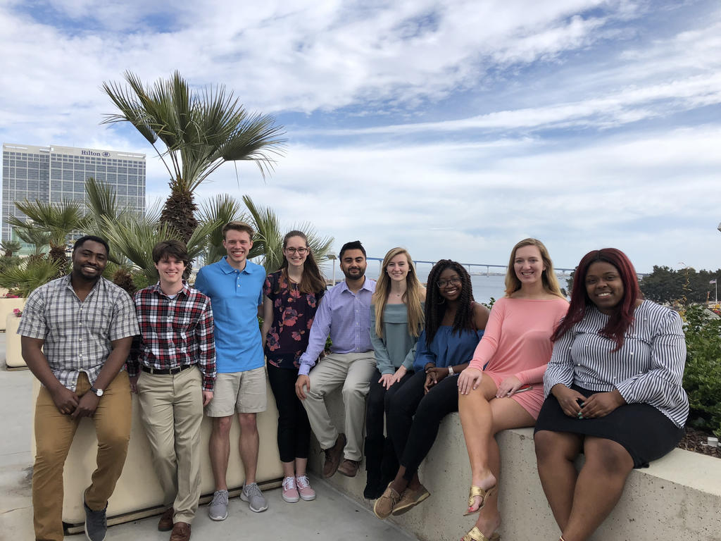 group shot of nine student scientists posing outside on a balcony; sky and ocean in the background