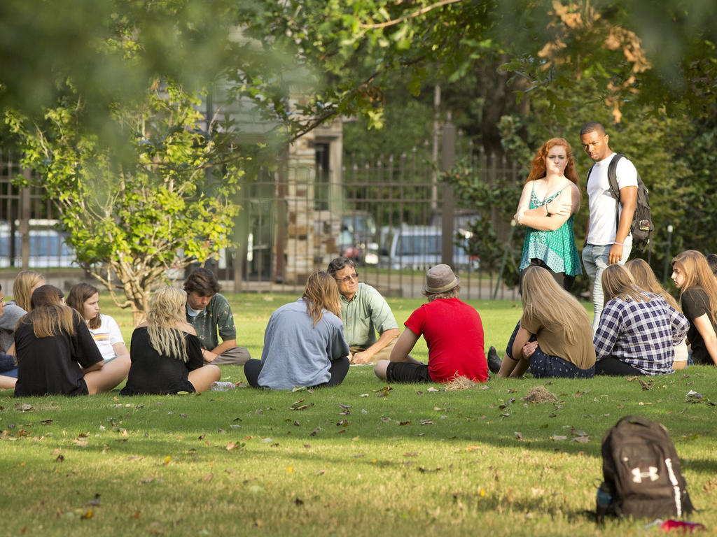 a diverse group of students and professors sitting in a circle outdoors in the shade under trees