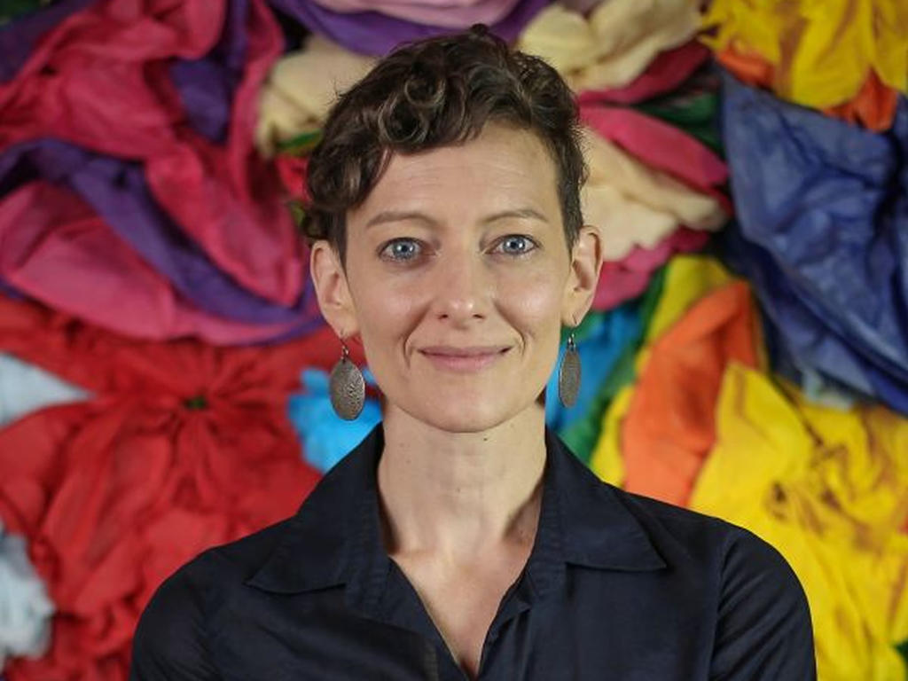 smiling professor in black blouse in front of a background made out of red, yellow, blue and orange cloth
