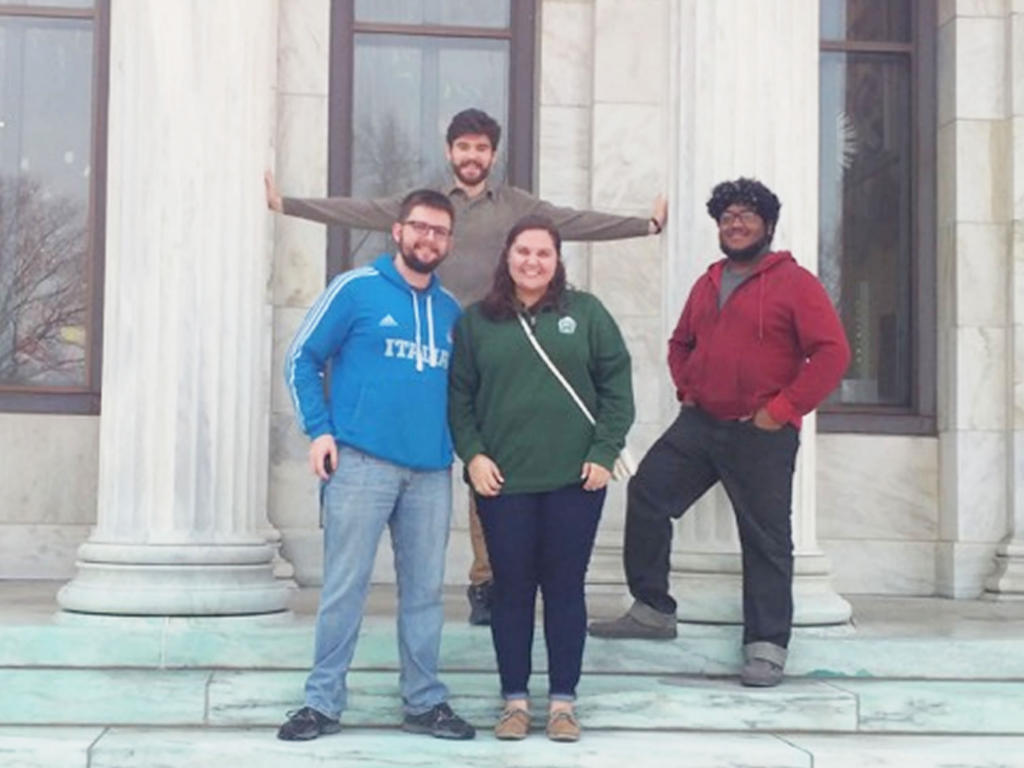 a diverse group of three males and one female student standing near large columns in casual attire