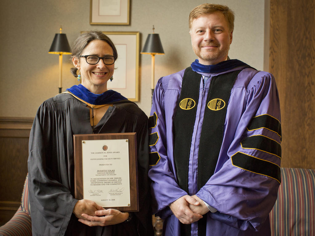 a female professor proudly holding her award next to the dean; both are sporting academic robes