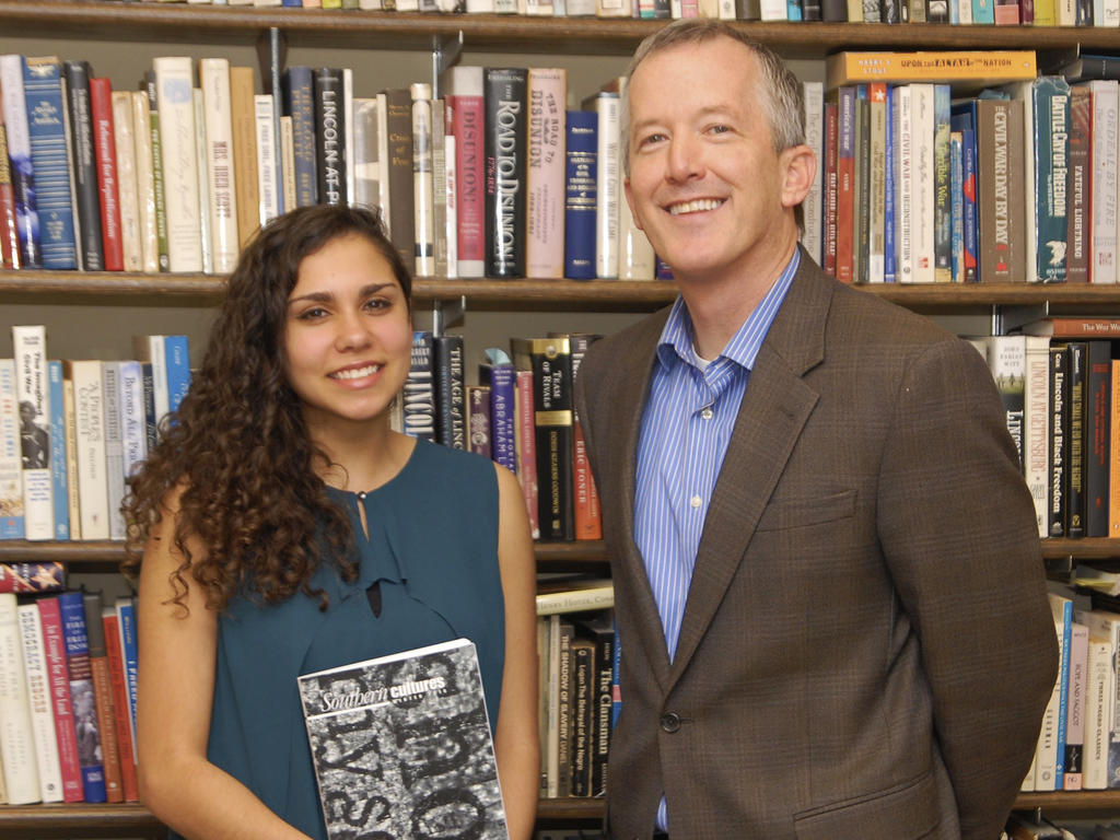a young woman proudly holding a history book while standing next to a white, male professor-- both standing in front of bookshelves 