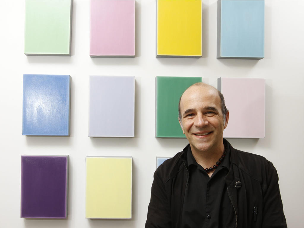 an older man standing in front of a wall collage of colorful canvases
