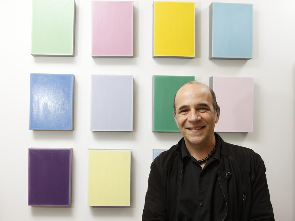 an older, male professor standing in front of a collection of monotone canvases hanging on a white wall