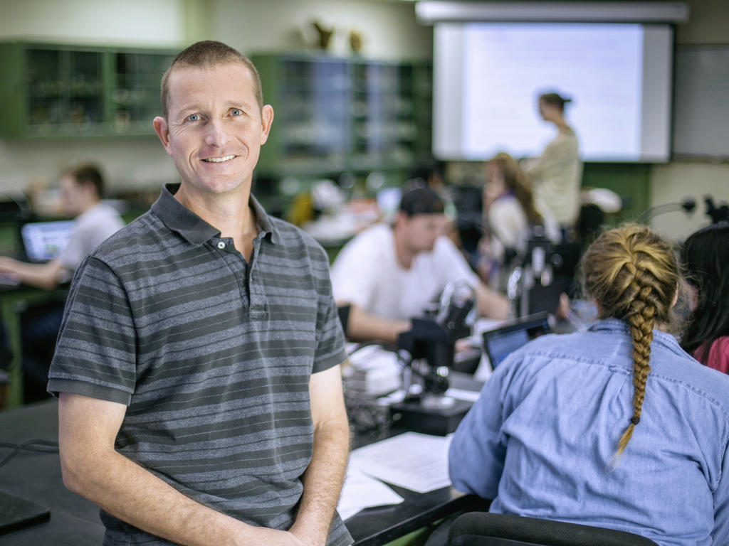 a middle-aged white male professor standing and smiling with students in a classroom behind him working