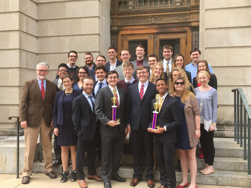 a diverse group of students wearing business professional attire and holding two trophies