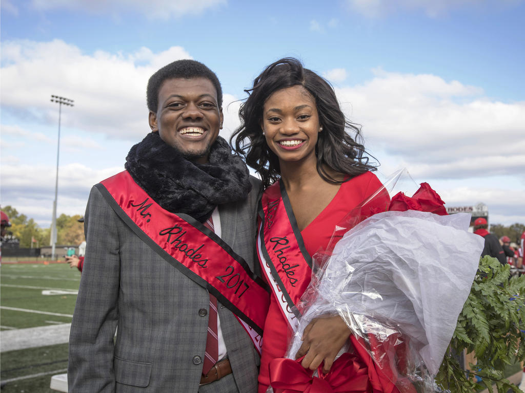 male and female college students, each draped in a red homecoming sash, standing on a football field