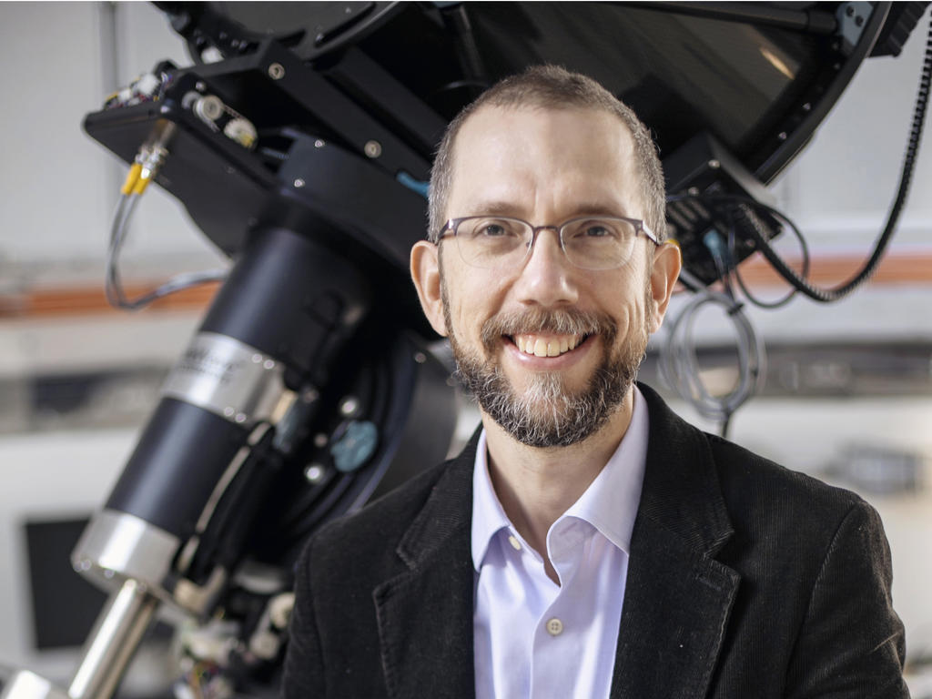 a middle-aged male professor standing and smiling in front of astronomy/physics equipment
