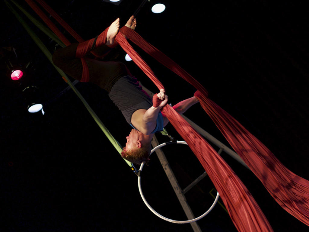 A muscular acrobat-man supporting his bodyweight using red aerial ropes while hanging upside down