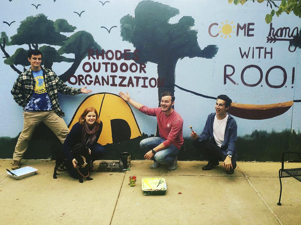 four diverse students sitting near the Rhodes Outdoor Organization's sign/logo