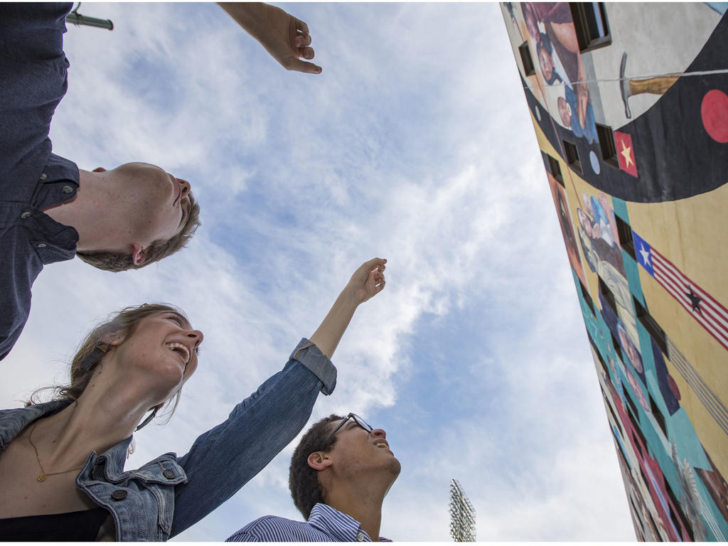 young adults pointing up at a mural on a building; a blue sky is the background