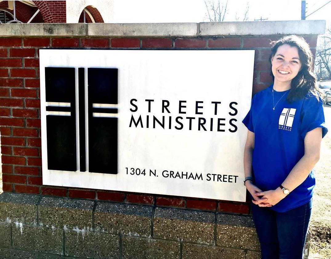 A woman in a blue t-shirt stands in front of a sign inlayed in brick that says Streets Ministries