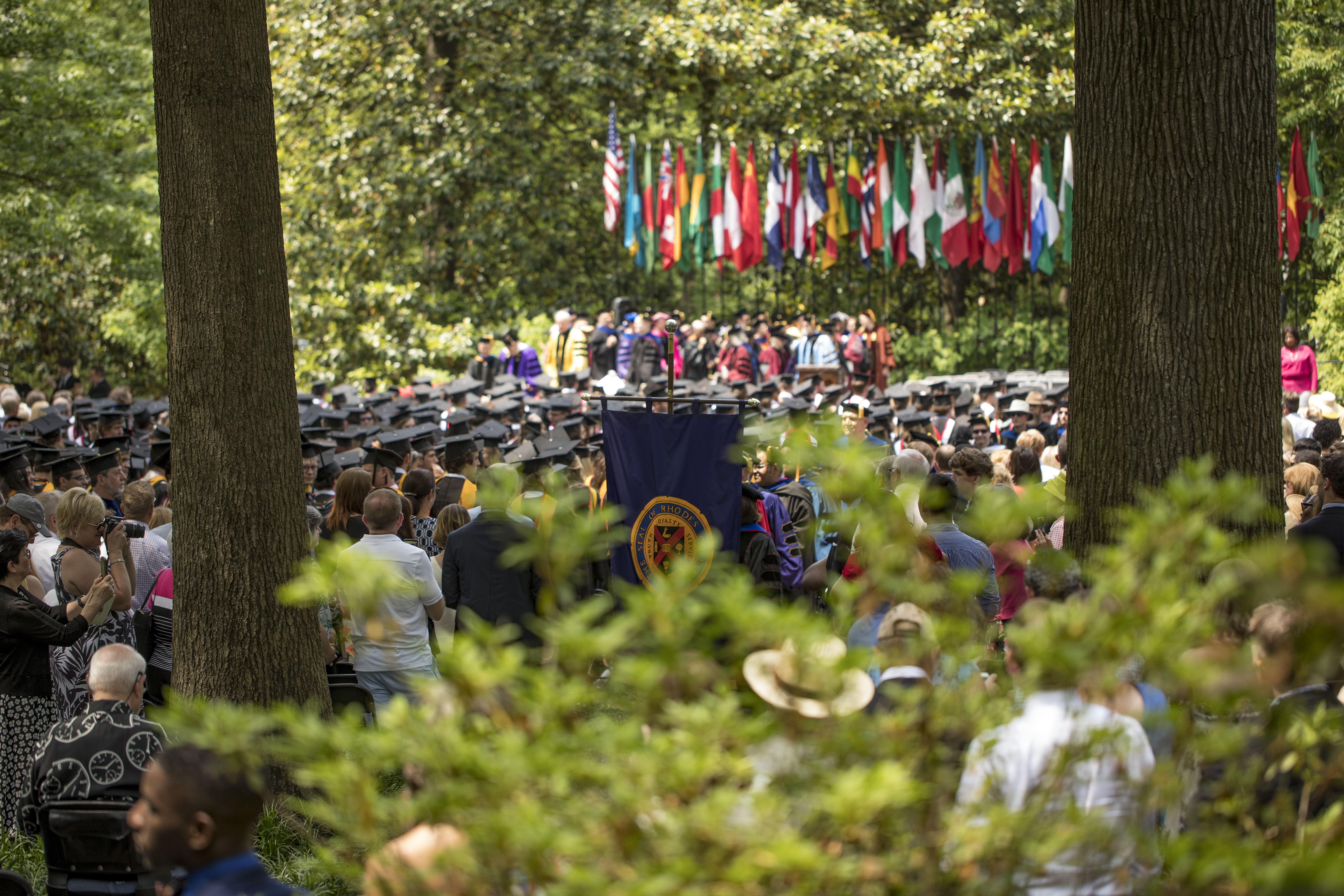 Crowd of graduates and families standing in front of a stage in a wooded area.