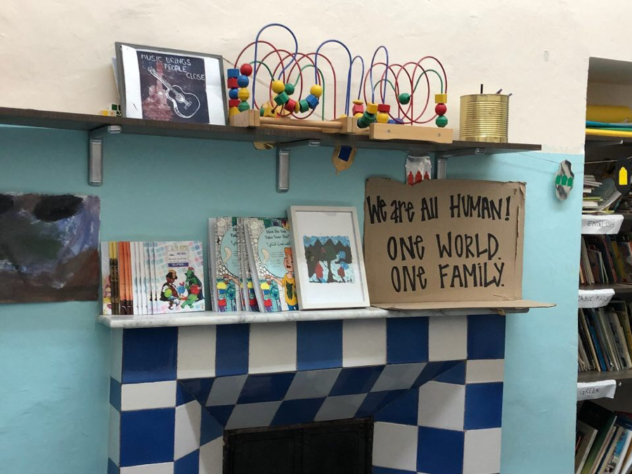 A shelf on a blue wall covered in books, toys, and a sign that reads: "We are all human. One world. One family."