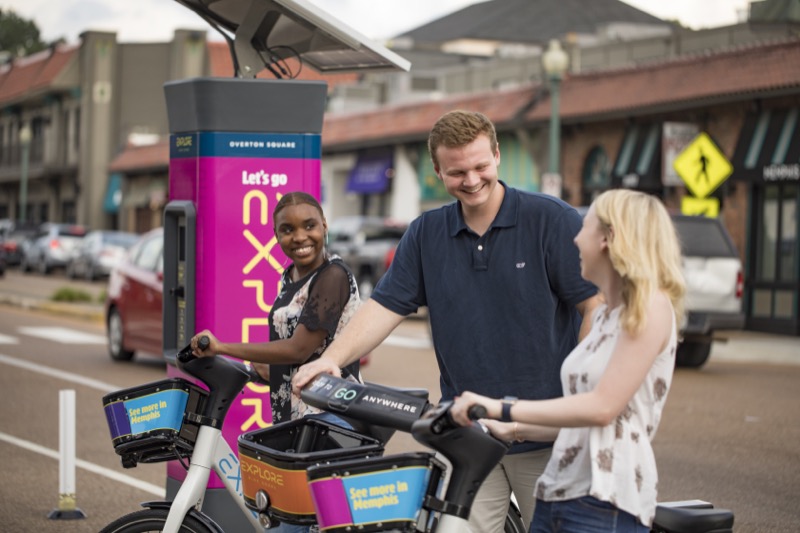 A young man and woman rent bikes from a rideshare station in a small shopping center.