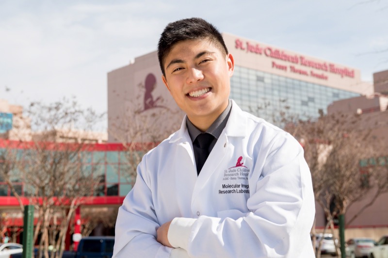 A young man in a lab coat stands in front of St Jude Children's Research Hospital.