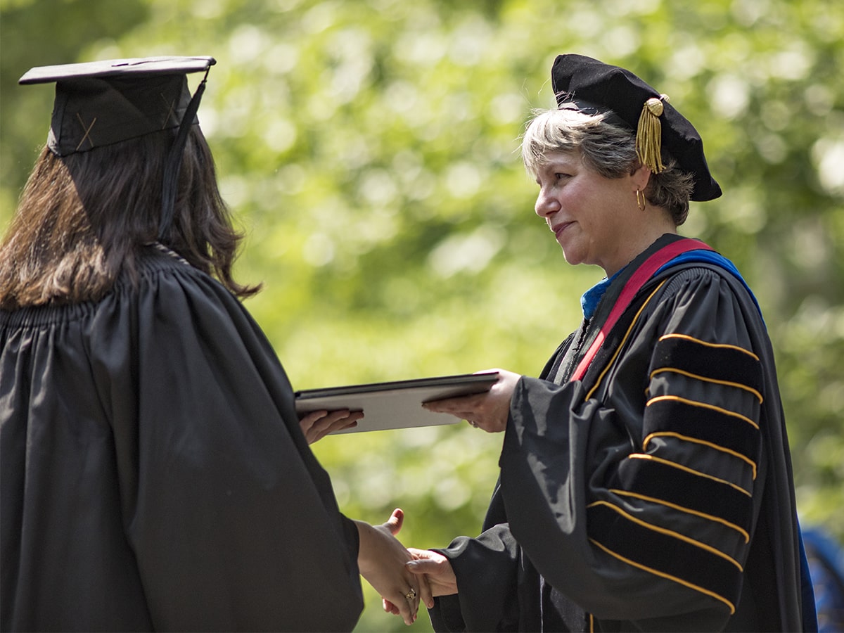President Hass in full regalia, handing a diploma to a student with long brown hair whose back is to the camera