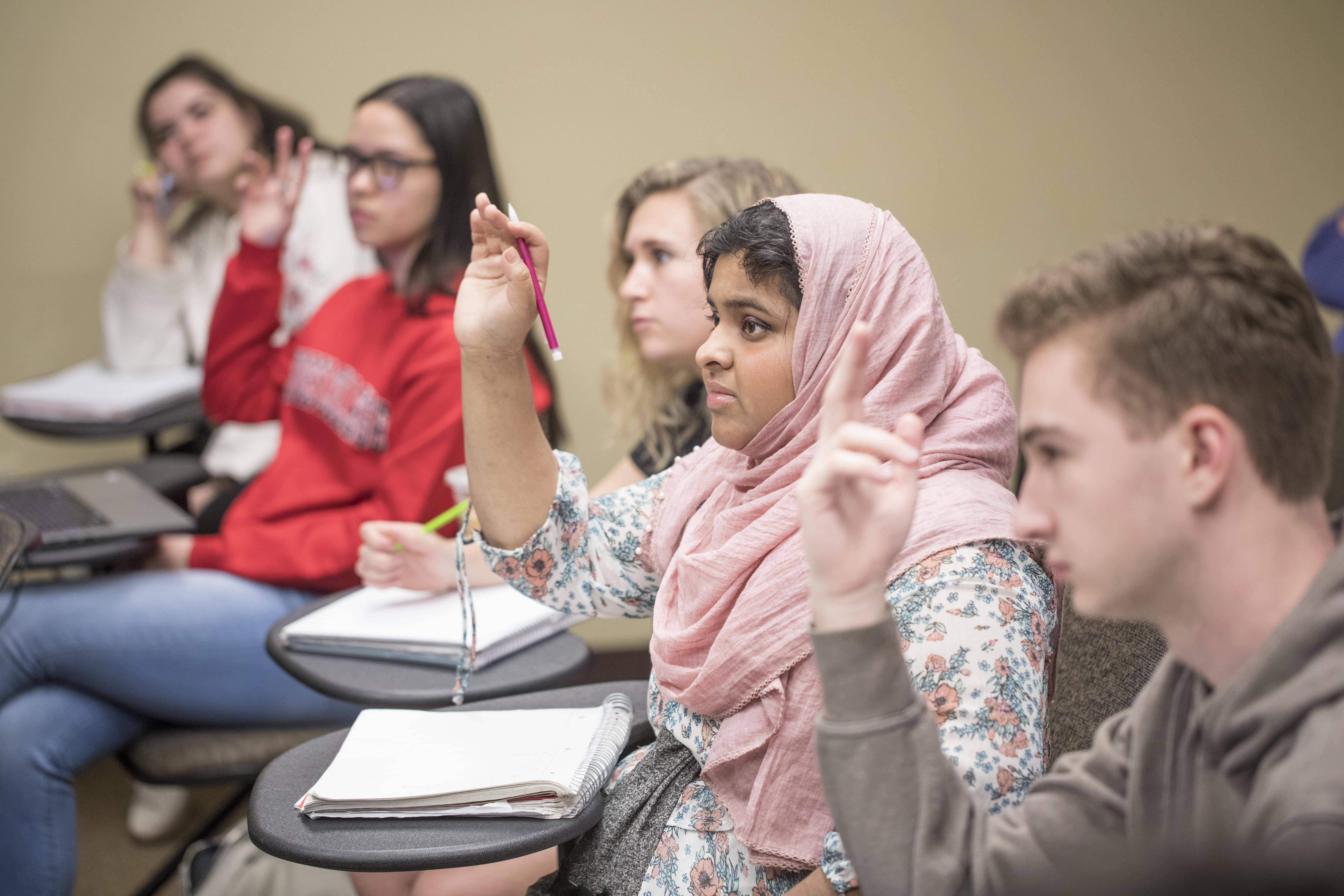 Five students are sitting at their desks, three with hands raised; the focus is on a young woman in a pink hijab