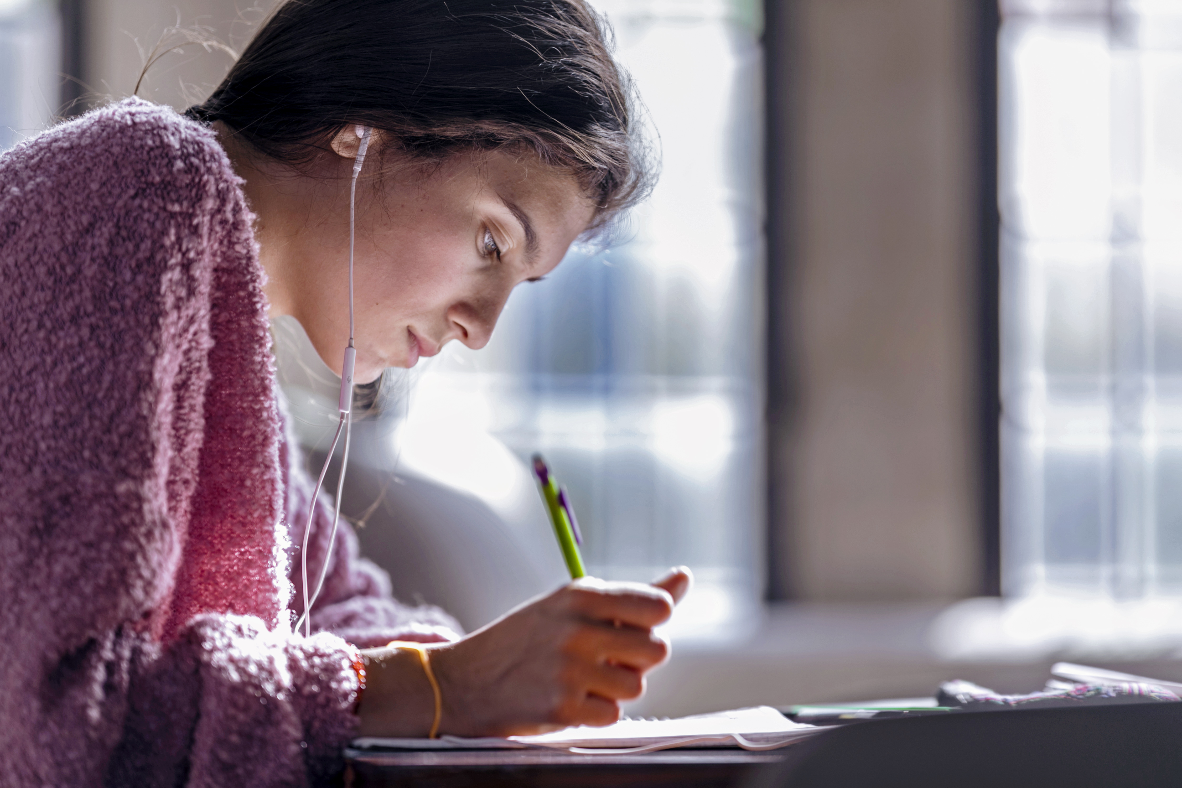 A young woman with dark hair and a pink sweater looks down while writing as light streams through the windows behind her. 