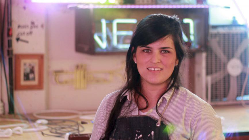 a young woman with dark hair stands in a studio in front of a neon sign