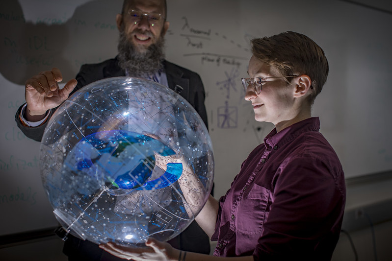A professor stands behind a clear model of the milky way. Light shines up from beneath the model, onto the face of a student to whom he is explaining it.