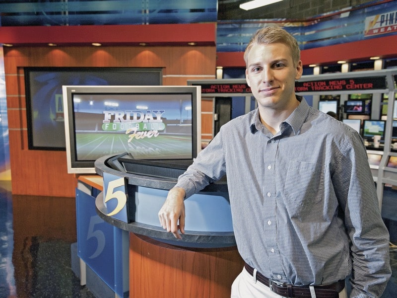 A man in an open-collared button down leans casually against the news desk and WMCTV.