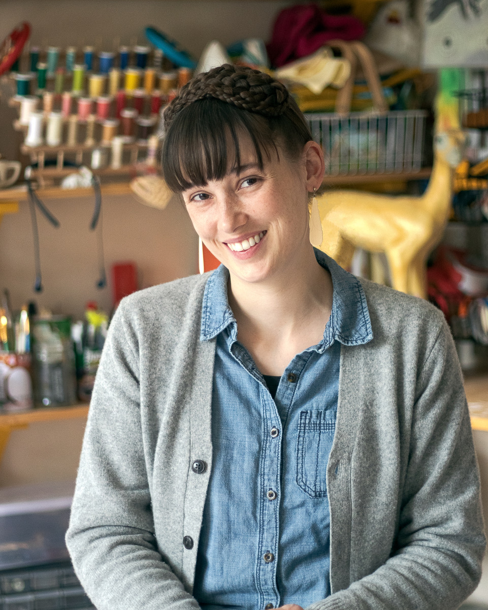 a female bangs and pulled back hair, wearing a sweater, stands before a wall of art objects 