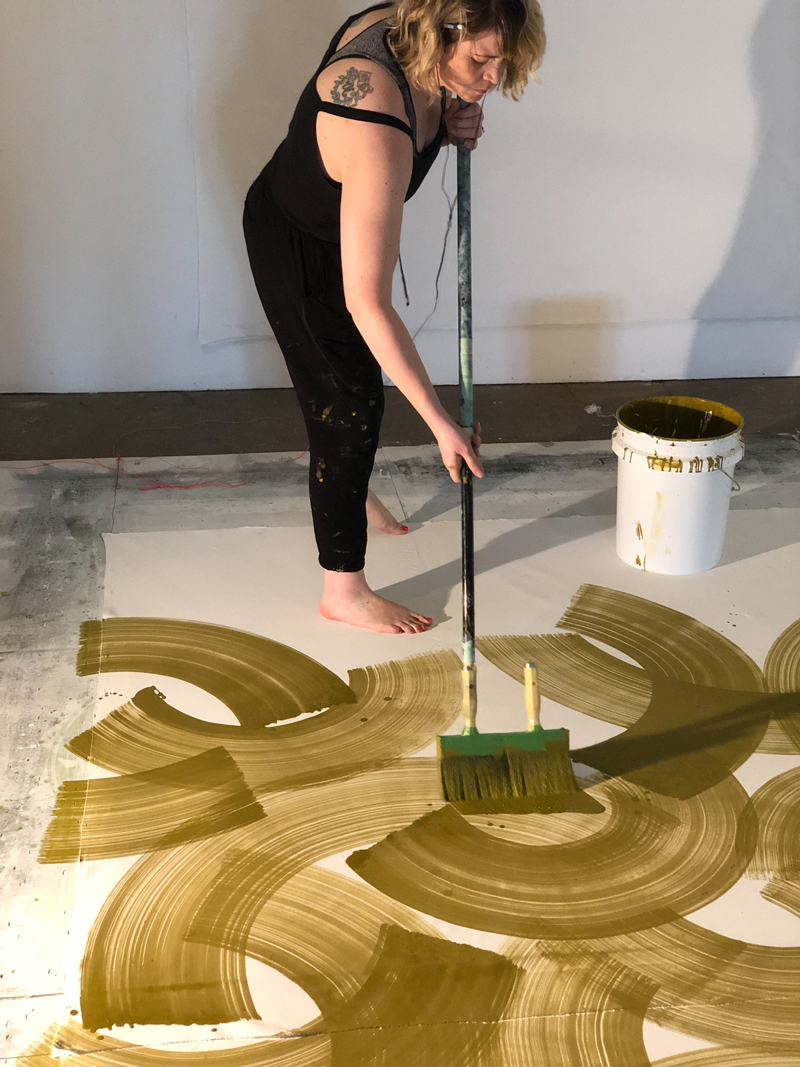 A woman dressed in black stands on a canvas and uses a large brush on a handle to paint an iimage