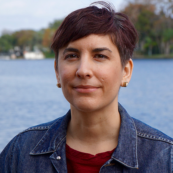 a woman with short dark hair in front of a lake smiles at the camera
