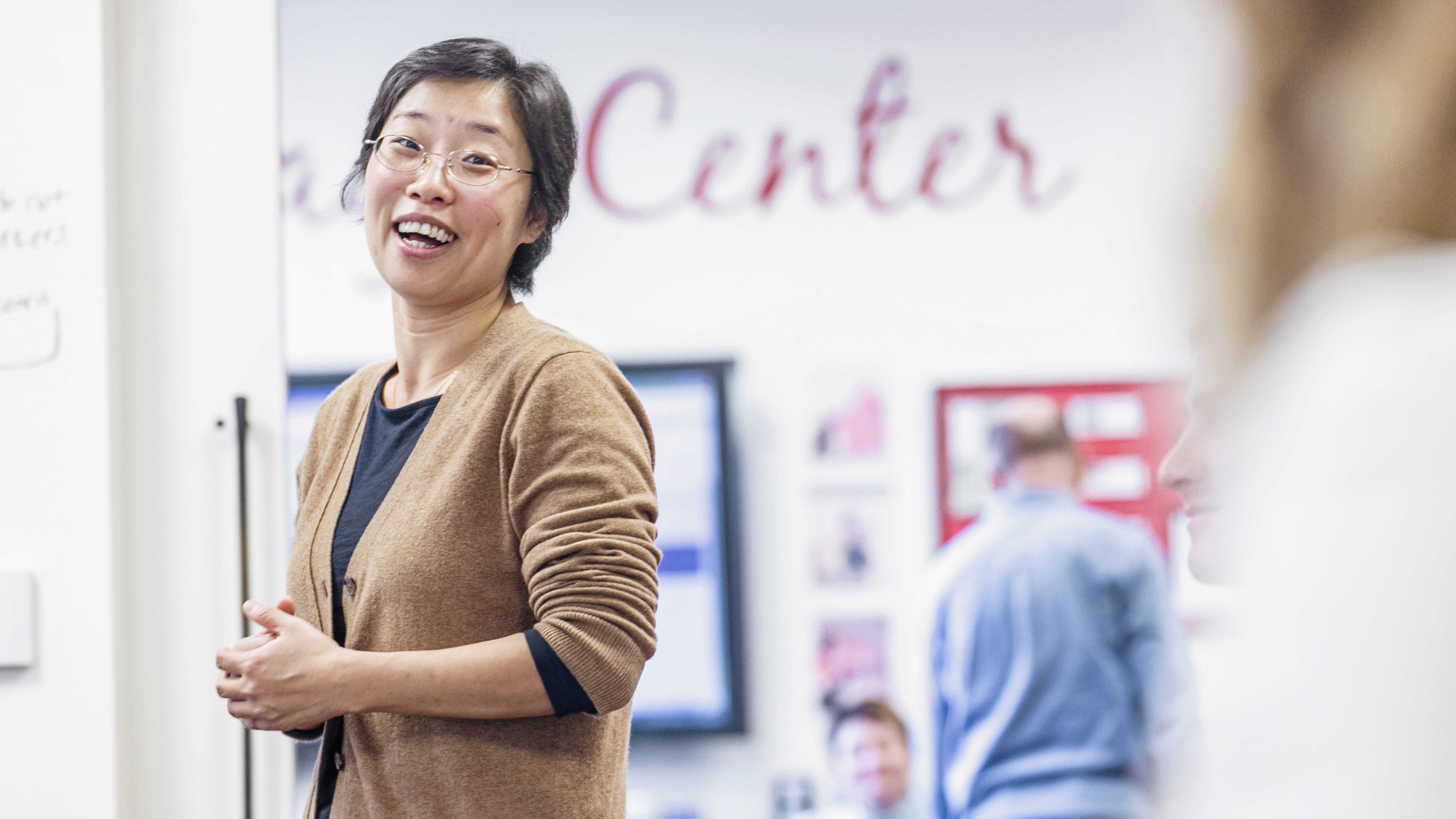 An Asian woman in a tan sweater laughs with students