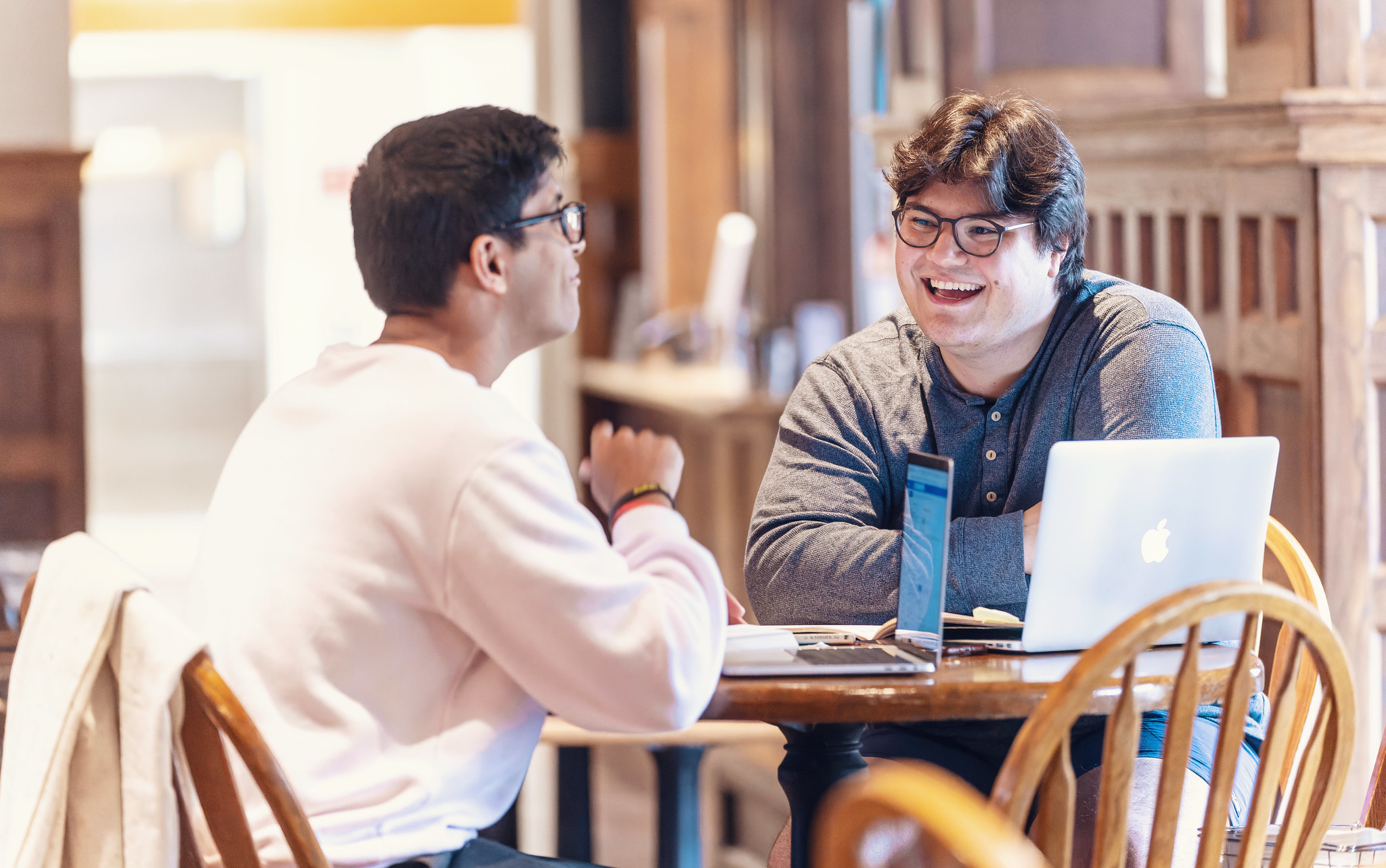 Two male student sit talking at a table. On the table is a laptop with several stickers on the back.