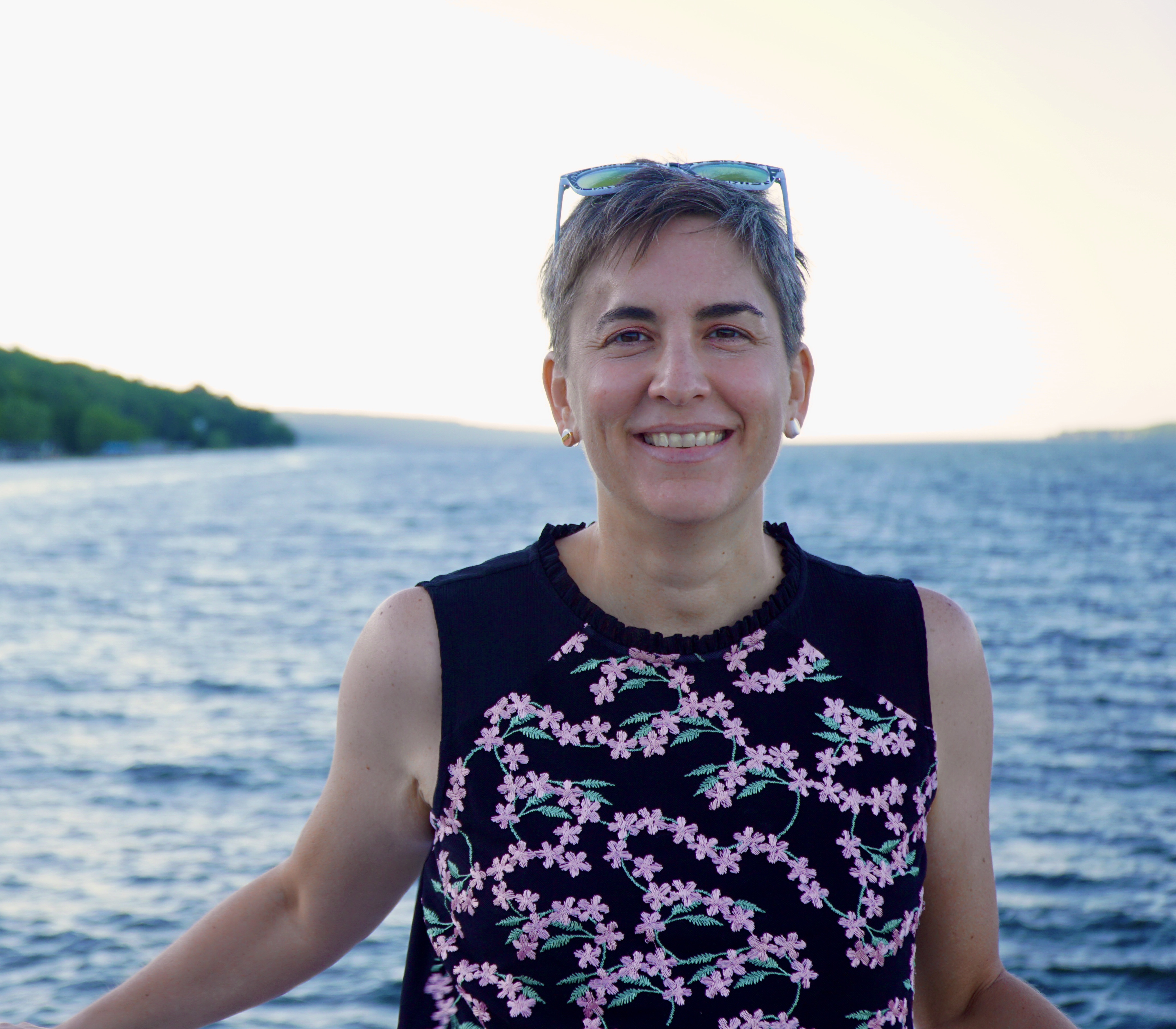 a woman with short dark hair in front of a lake smiles at the camera