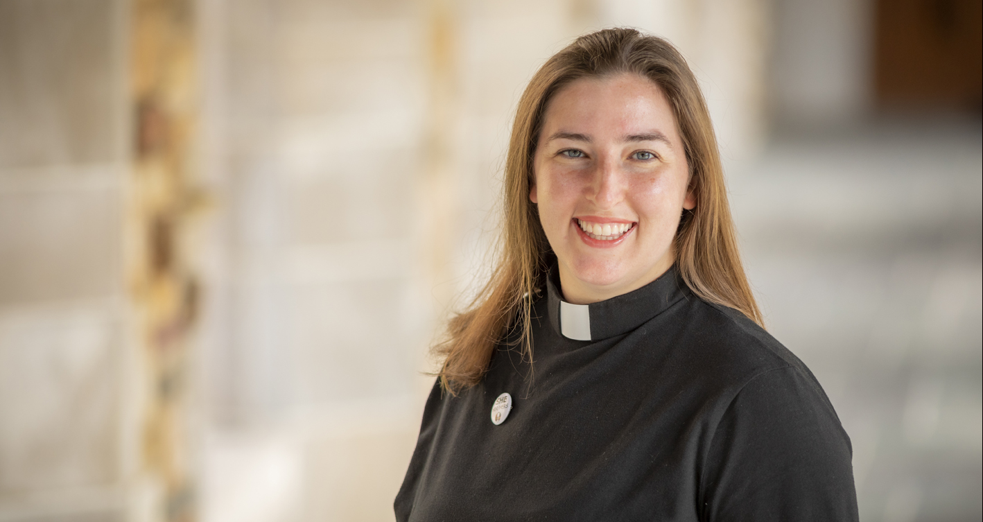 A young, dark haired woman in clerical vestments smiles at the camera