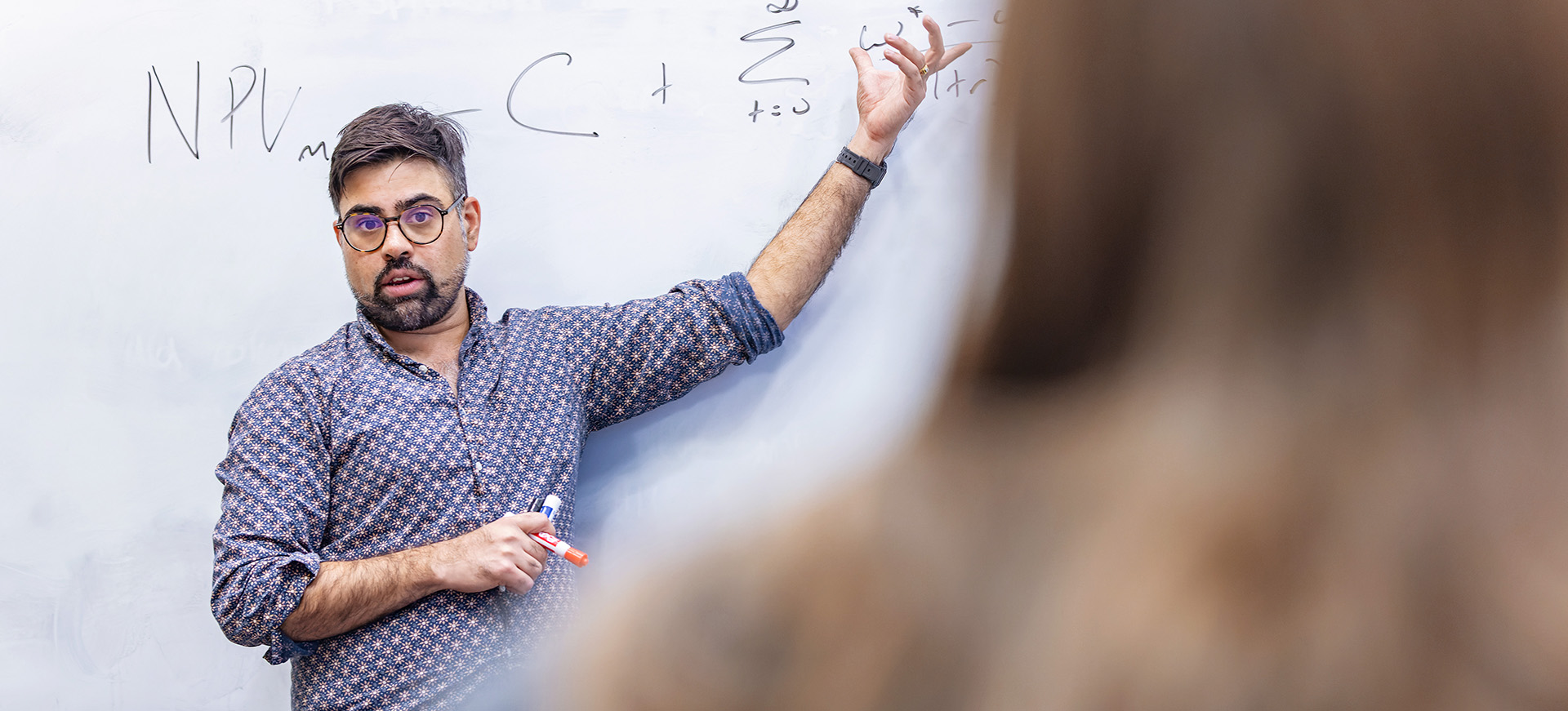 a male professor gestures at a whiteboard with equations