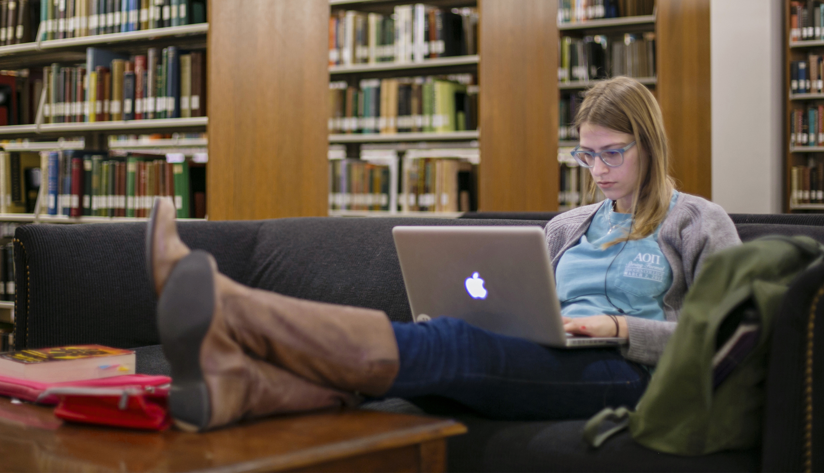 A student sits with her feet up on the coffee table, a laptop on her lap; she is among shelves of books