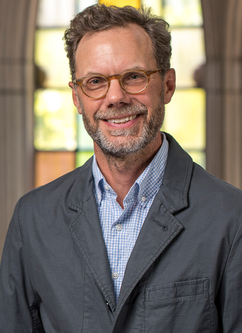 a man with a short beard, moustache, and glasses in front of a stained glass window