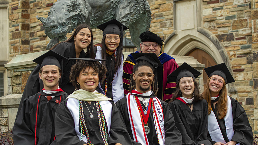a group of students in graduatoin robes pose by a statue of a lynx