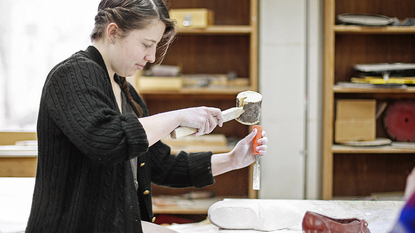 A woman in a dark jacket uses a large wooden mallet and a chisel to knock away at a granite carving of a shoe.