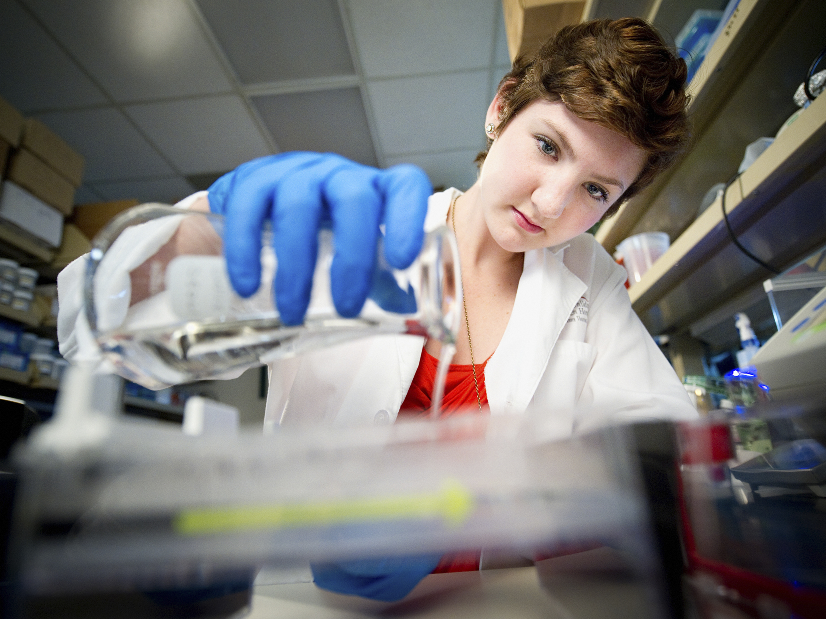 A short haired young woman in a lab coat and blue latex gloves pours liquid into a beaker.