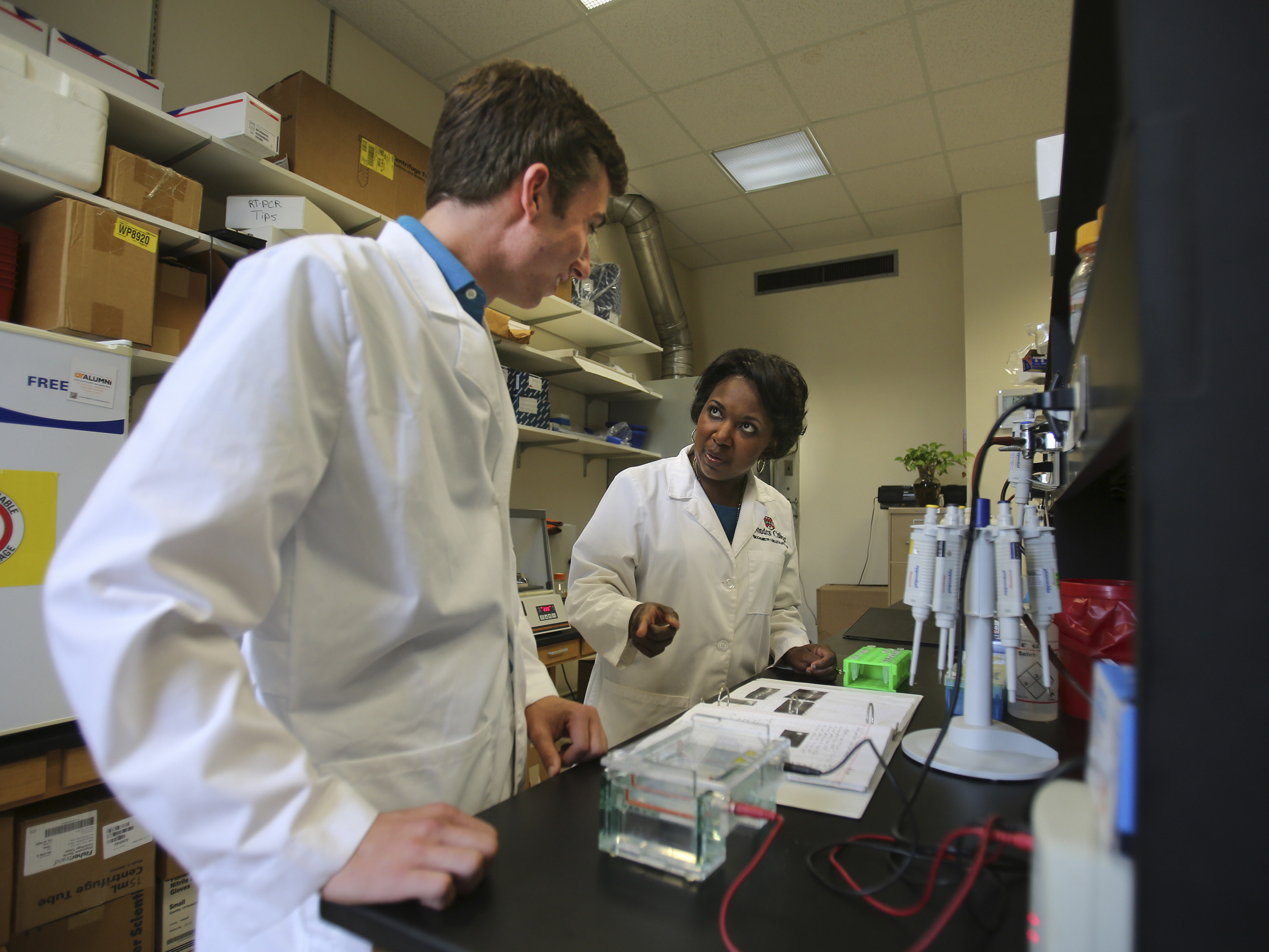 Two researchers in white coats stand at a lab table reviewing notes.