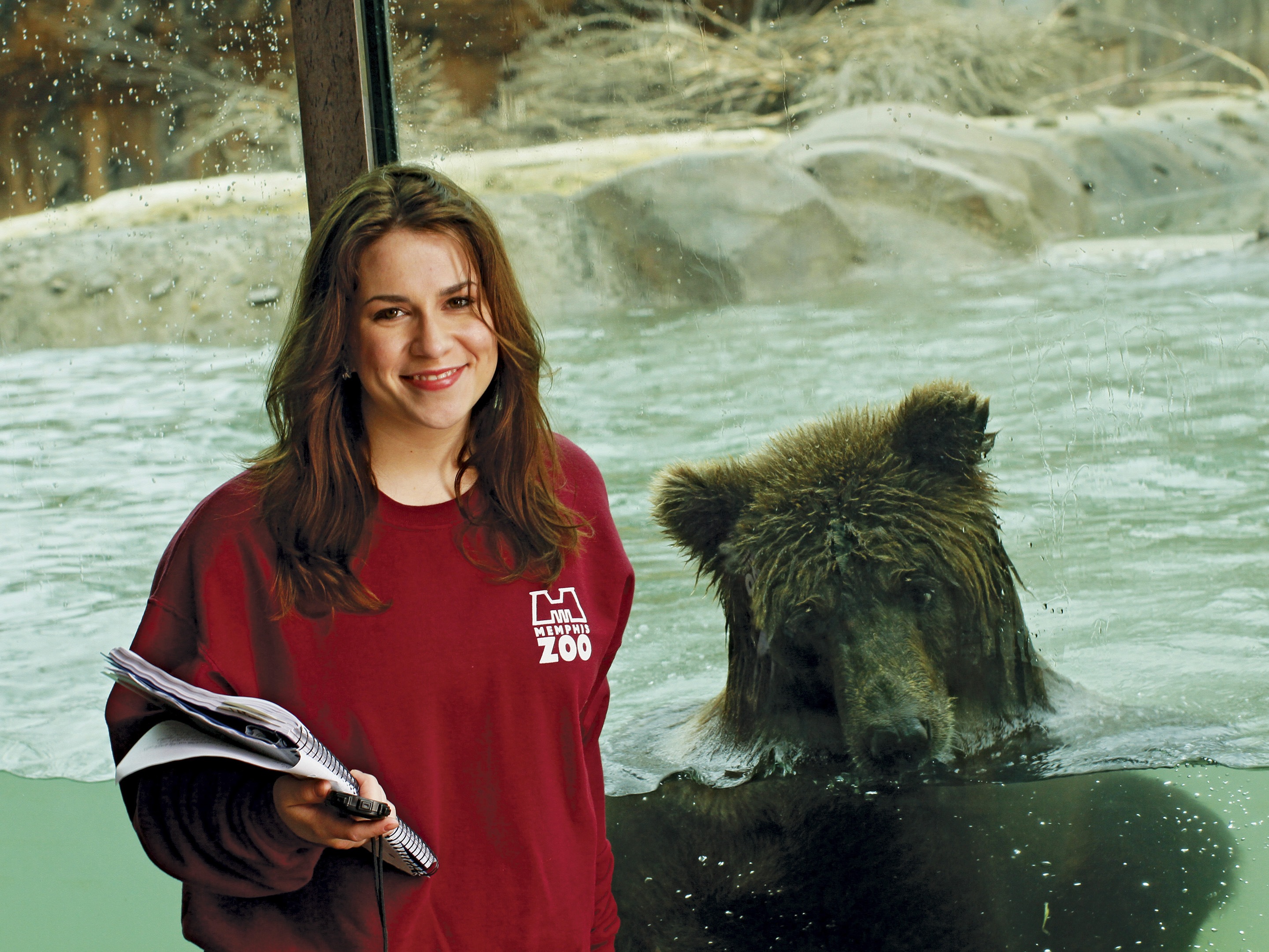 A young woman with a clipboard stands in front of a grizzly bear.