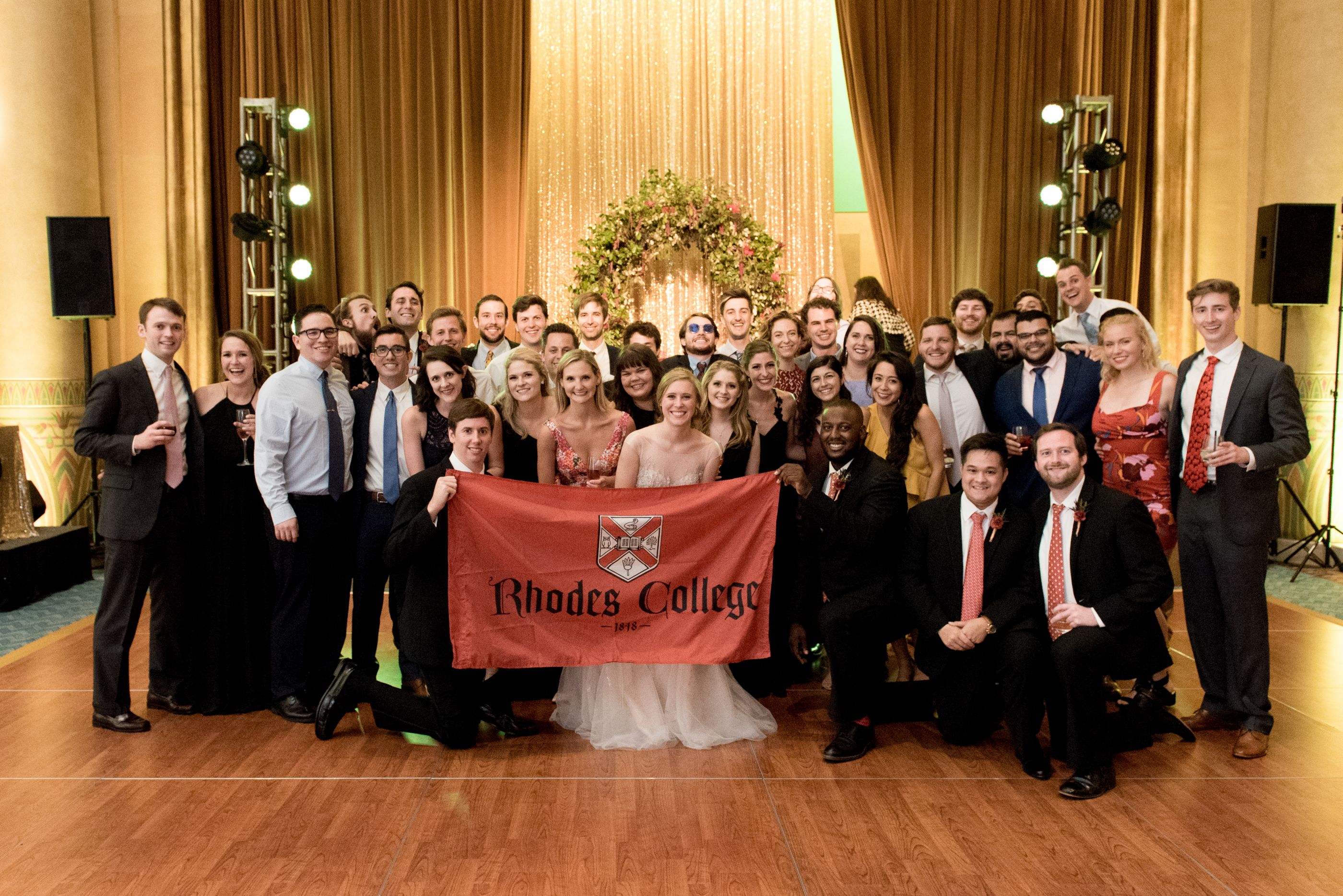 A large group of young Rhodes alumni and students holding a Rhodes flag. The person in the center is wearing a wedding dress. 