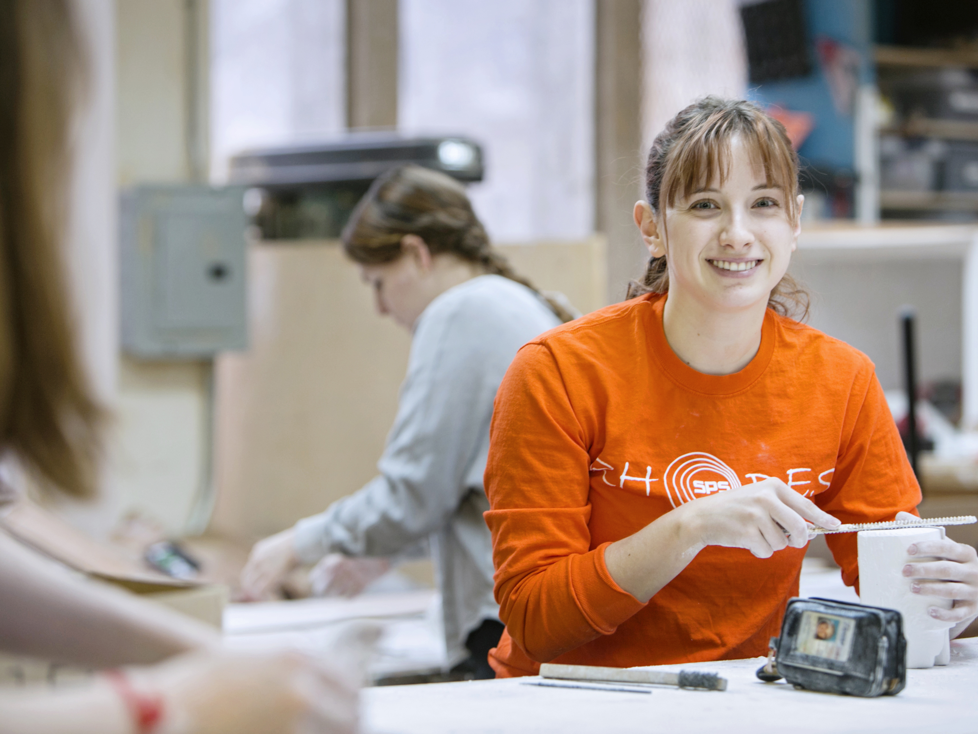 A brown haired young woman in an orange sweater smiles for the camera.