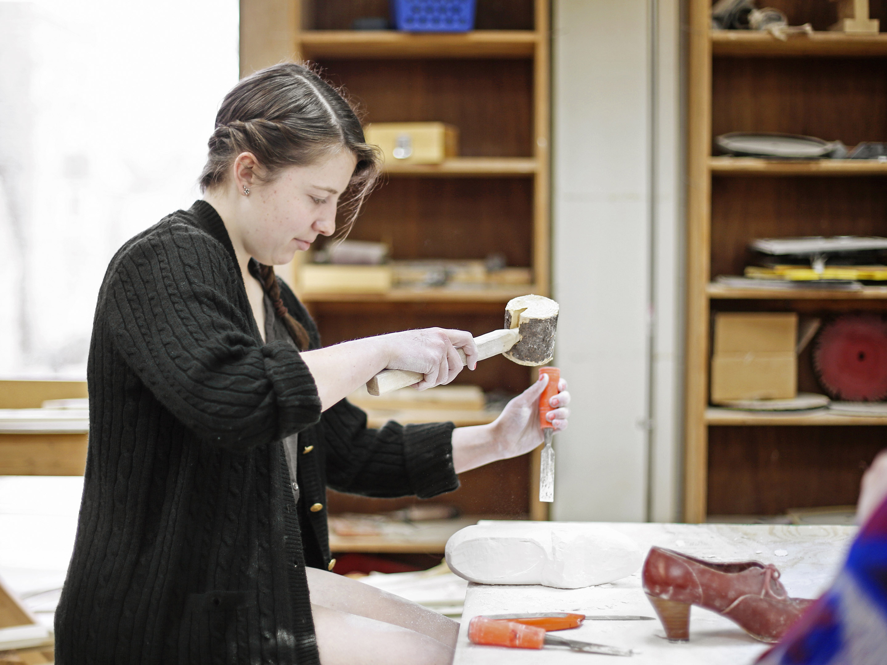 A woman in a dark jacket uses a large wooden mallet and an orange chisel to knock away at a granite carving of a shoe.