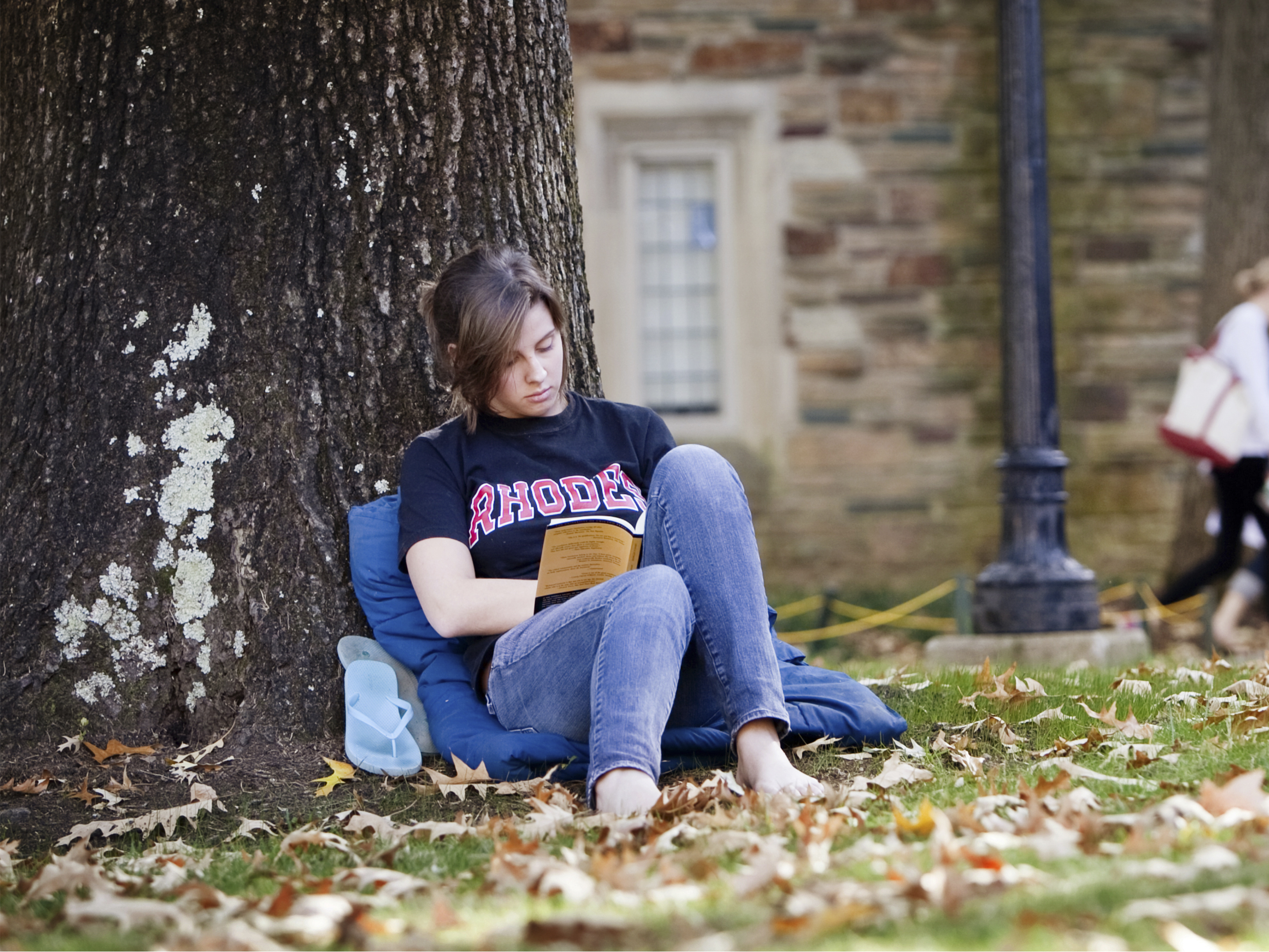 A barefoot young woman in a Rhodes tshirt and jeans sits leaning against a tree reading a book.