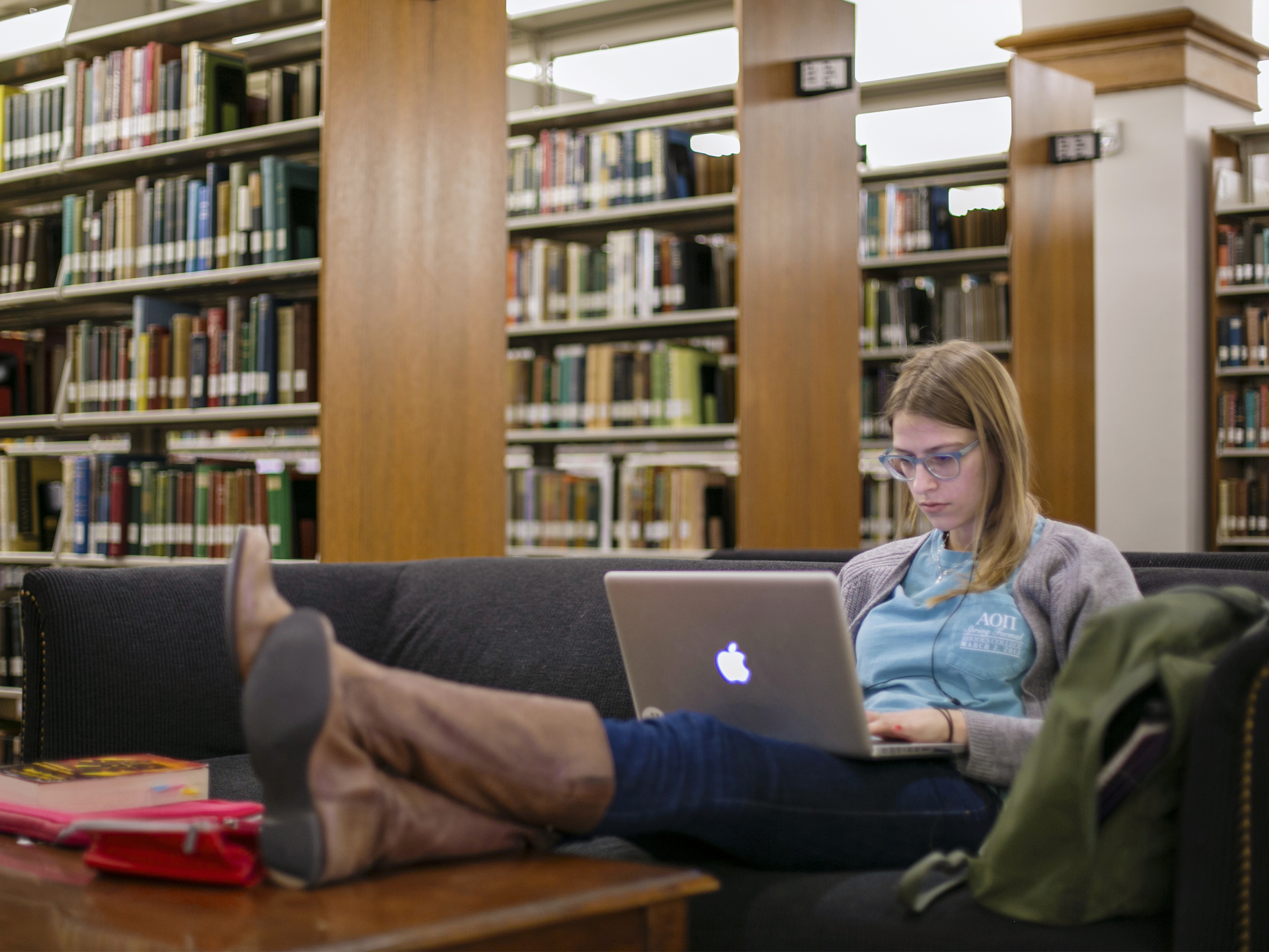 A student sits with her feet up on the coffee table, a laptop on her lap; she is among shelves of books