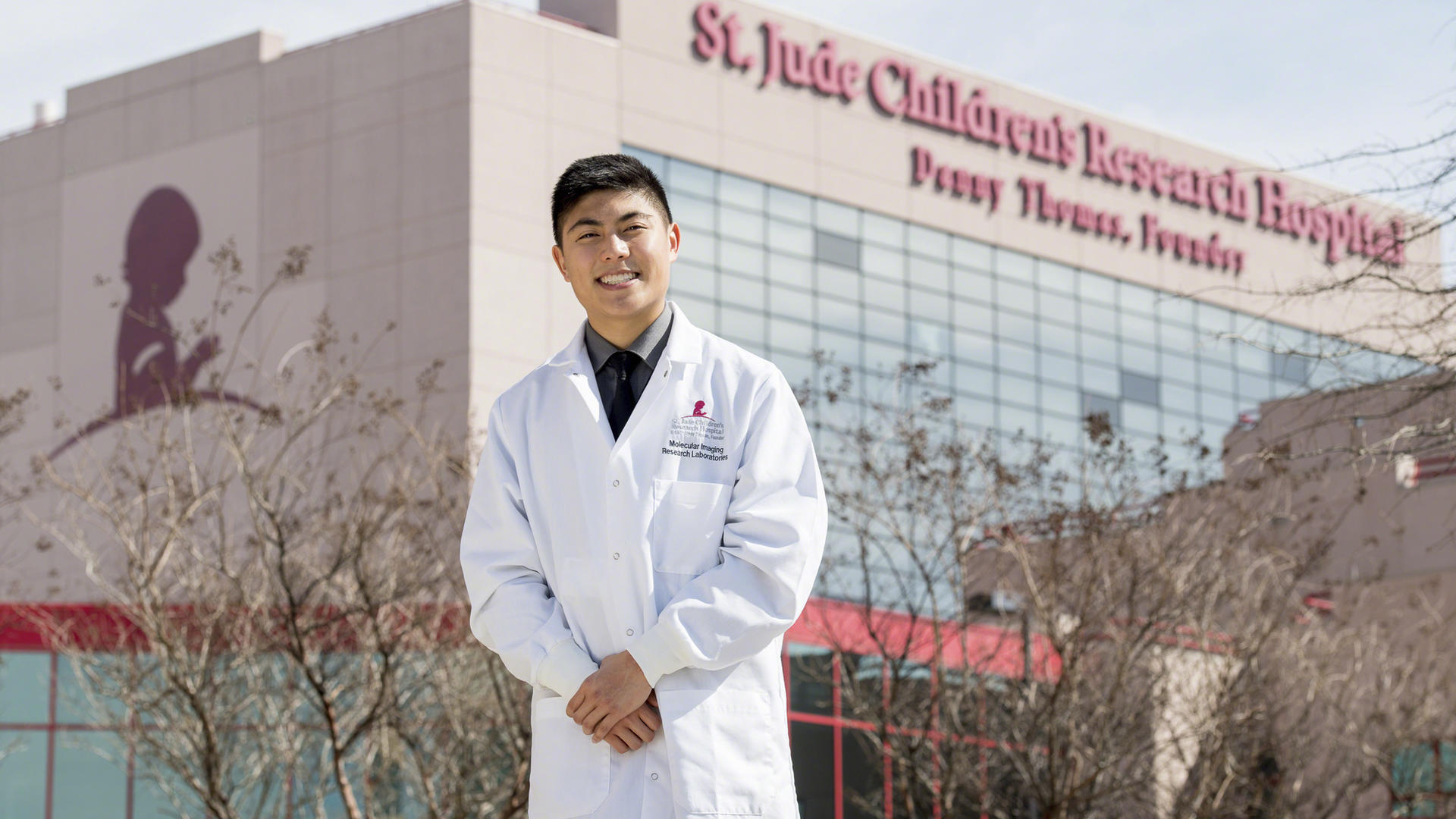 A young man in a white coat stands outside St. Jude Children's Research Hospital.