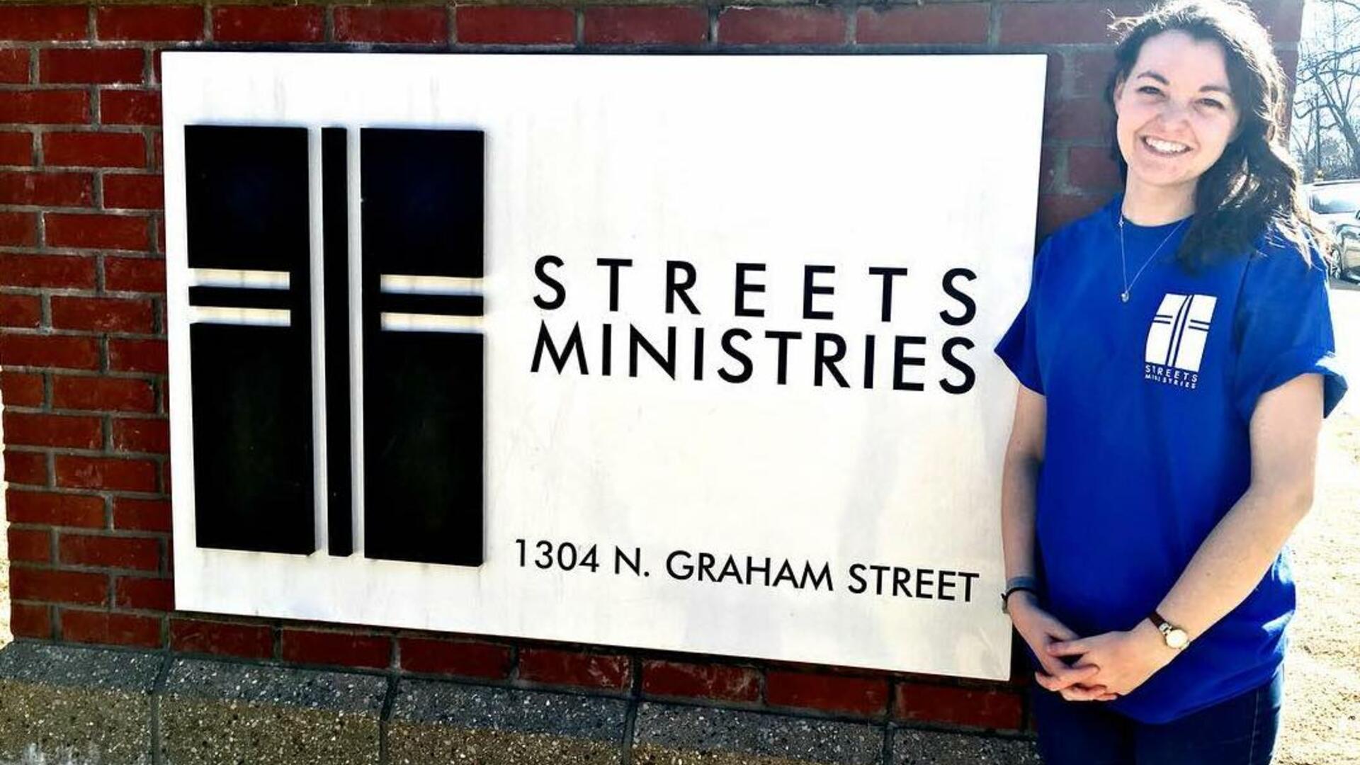 A woman in a blue t-shirt stands in front of a sign inlayed in brick that says Streets Ministries